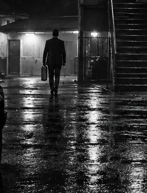 A figure with a briefcase walks through a rain-slicked courtyard at night.